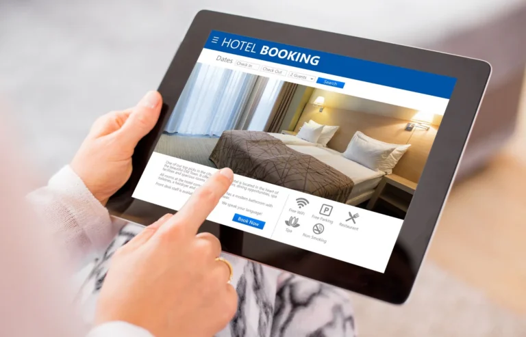 Ένα άτομο που χρησιμοποιεί ένα tablet για να περιηγηθεί σε έναν ιστότοπο κρατήσεων ξενοδοχείων που έχει σχεδιαστεί για άτομα με ειδικές ανάγκες, εστιάζοντας στην επιλογή δωματίου και στην προβολή των ανέσεων που προσφέρει το ξενοδοχείο.